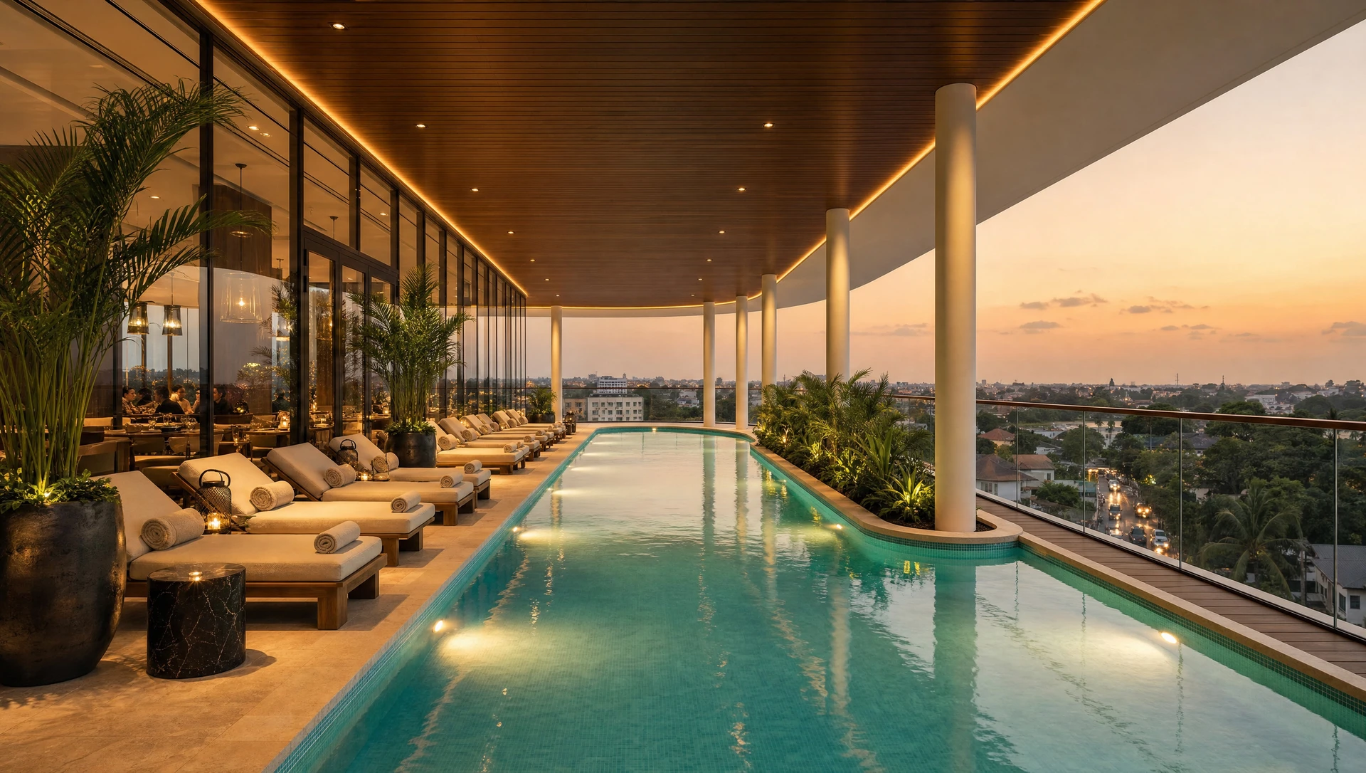 The pool at dusk, loungers and planting along one side beneath a timber soffit, the city beyond the glass